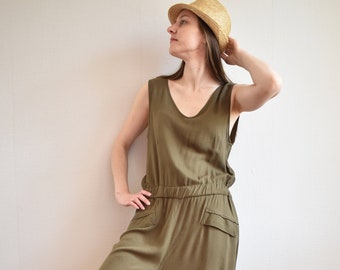 May include: A woman wearing a green jumpsuit with a straw hat and white sneakers. The jumpsuit has a wide leg and a v-neck.