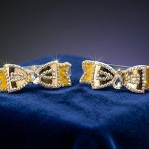 May include: Two gold-toned bow hair clips with yellow accents and clear crystal embellishments. The bows are set on clear clips and rest on a blue velvet surface. The crystals sparkle, adding a touch of glamour.