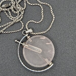 May include: A silver chain necklace with a round pendant featuring a clear pink crystal with a silver tube inside. The pendant is attached to a silver bar that rotates.