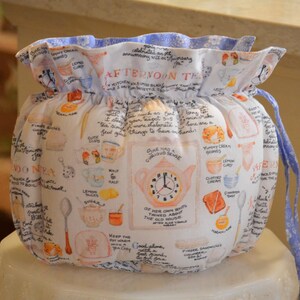 Może przedstawiać: Biała, materiałowa osłona na herbatę ze ściągaczem. Osłona ma fantazyjny wz&oacute;r filiżanek, imbryk&oacute;w i odręcznego tekstu. G&oacute;rna krawędź jest marszczona z niebieskim kwiatowym wzorem.