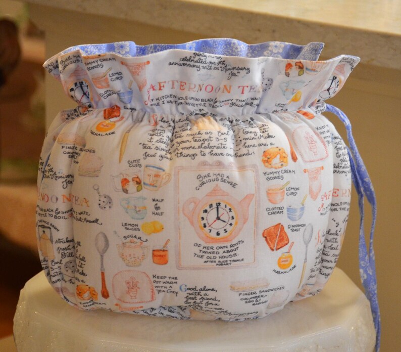 Może przedstawiać: Biała, materiałowa osłona na herbatę ze ściągaczem. Osłona ma fantazyjny wz&oacute;r filiżanek, imbryk&oacute;w i odręcznego tekstu. G&oacute;rna krawędź jest marszczona z niebieskim kwiatowym wzorem.