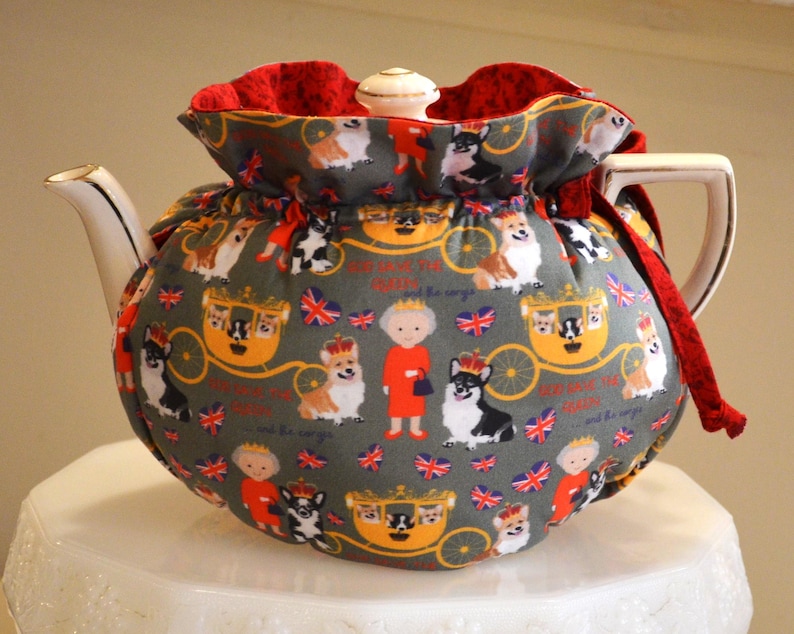 Puede incluir: Una tetera de cer&aacute;mica con una funda estampada con corgis, coronas y un carruaje real sobre un fondo gris. La funda tiene un interior rojo y un asa y boquilla blancos. La tetera est&aacute; sobre una superficie blanca.