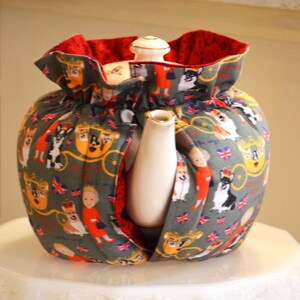 Puede incluir: Un calentador de t&eacute; gris con interior rojo, con un estampado de corgis, figuras reales y carruajes. El calentador envuelve una tetera blanca con boquilla y tapa doradas.