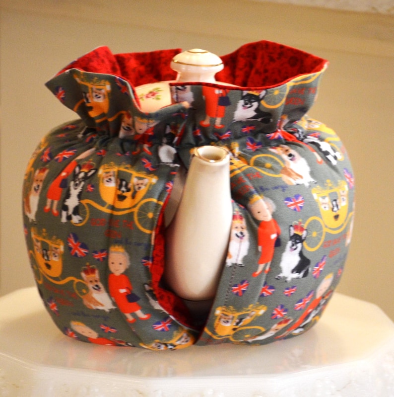 Puede incluir: Un calentador de t&eacute; gris con interior rojo, con un estampado de corgis, figuras reales y carruajes. El calentador envuelve una tetera blanca con boquilla y tapa doradas.