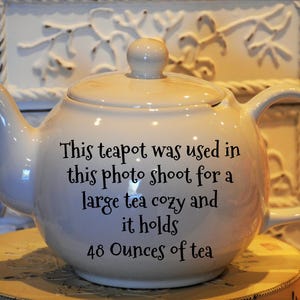 May include: Cream-colored ceramic teapot with a lid and handle. The teapot has text that reads, "This teapot was used in this photo shoot for a large tea cozy and it holds 48 Ounces of tea."