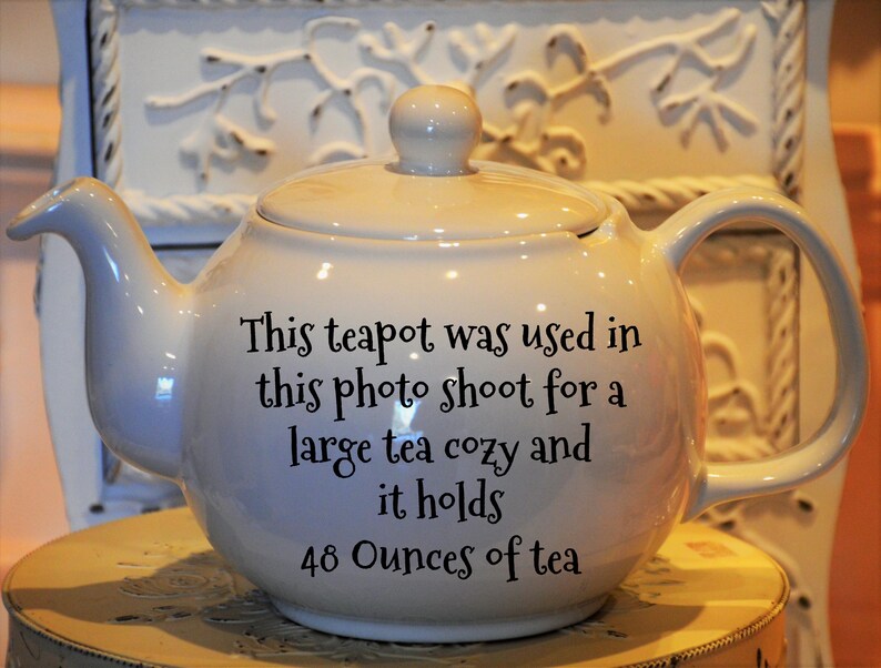 May include: Cream-colored ceramic teapot with a lid and handle. The teapot has text that reads, "This teapot was used in this photo shoot for a large tea cozy and it holds 48 Ounces of tea."