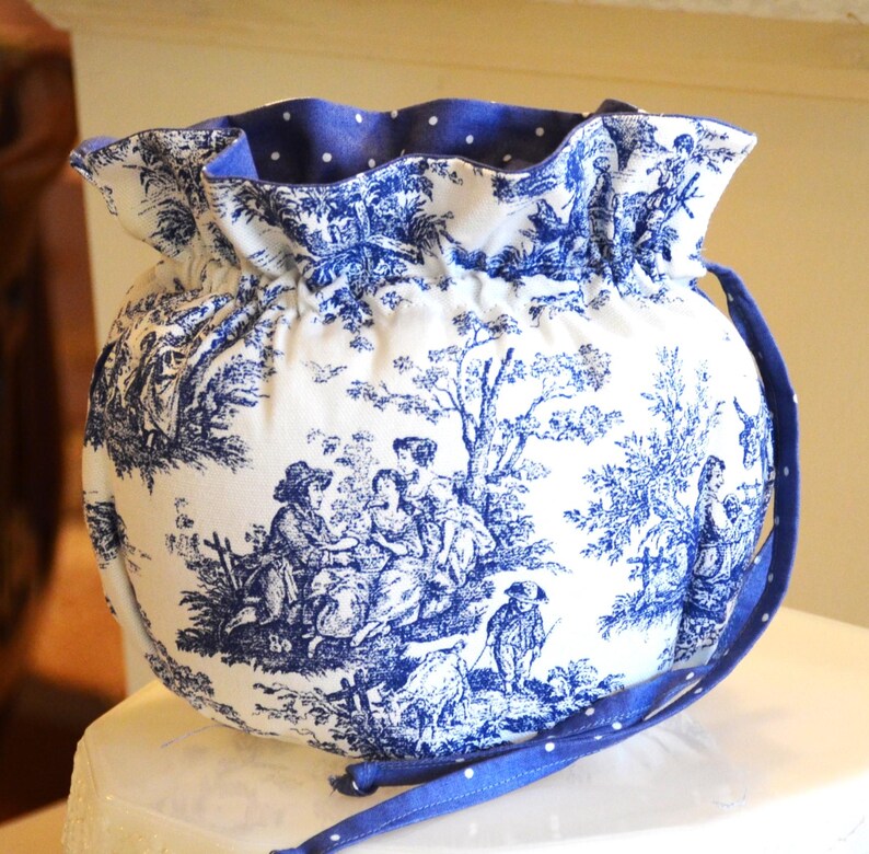 Op de afbeelding: Een ronde trekkoordtas in een blauw en wit toile de Jouy-ontwerp. De tas heeft een klassieke blauw-witte pastorale sc&egrave;neprint. De binnenkant en het trekkoord zijn effen blauw met witte stippen. De tas heeft een blauwe band.