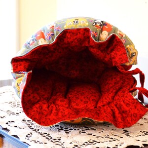 Puede incluir: Una bolsa redonda de tela, abierta, con interior rojo y cierre de cord&oacute;n. El exterior presenta un estampado de guardias brit&aacute;nicos, banderas y dise&ntilde;os tem&aacute;ticos de Londres. La bolsa est&aacute; sobre una superficie de encaje blanco.