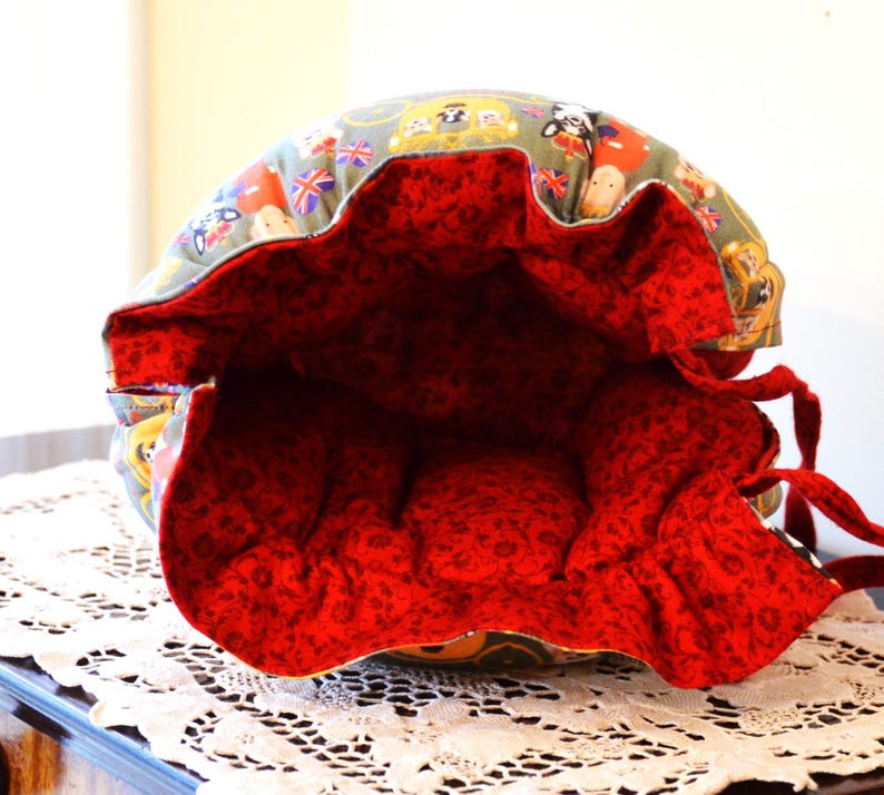 Puede incluir: Una bolsa redonda de tela, abierta, con interior rojo y cierre de cord&oacute;n. El exterior presenta un estampado de guardias brit&aacute;nicos, banderas y dise&ntilde;os tem&aacute;ticos de Londres. La bolsa est&aacute; sobre una superficie de encaje blanco.