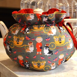 Puede incluir: Un calentador de t&eacute; gris con interior rojo, con un estampado repetido de corgis, coronas, carrozas y la bandera brit&aacute;nica. El calentador de t&eacute; envuelve una tetera blanca.