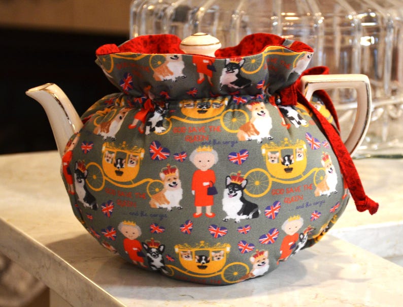 Puede incluir: Un calentador de t&eacute; gris con interior rojo, con un estampado repetido de corgis, coronas, carrozas y la bandera brit&aacute;nica. El calentador de t&eacute; envuelve una tetera blanca.