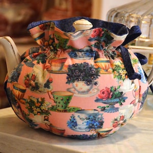 Puede incluir: Un calentador de t&eacute; floral con fondo rosa y un estampado de tazas de t&eacute; y flores. El calentador tiene un ribete azul marino y un cierre de cord&oacute;n. La tetera es blanca con un asa dorada.