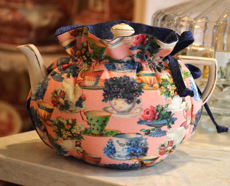 Puede incluir: Un calentador de t&eacute; floral con fondo rosa y un estampado de tazas de t&eacute; y flores. El calentador tiene un ribete azul marino y un cierre de cord&oacute;n. La tetera es blanca con un asa dorada.