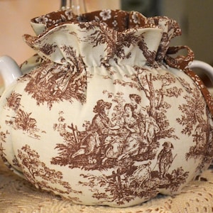 May include: A white ceramic teapot with a brown toile fabric cozy. The cozy features a pastoral scene with figures and trees. The lid and handle are white. The cozy has a ruffled edge and a drawstring closure.