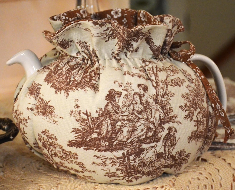 May include: A white ceramic teapot with a brown toile fabric cozy. The cozy features a pastoral scene with figures and trees. The lid and handle are white. The cozy has a ruffled edge and a drawstring closure.
