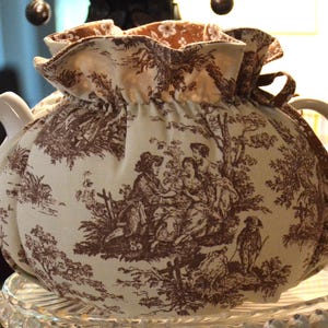 May include: A cream-colored teapot with a brown toile fabric cozy. The cozy features a pastoral scene with figures and trees. The teapot has a white handle and spout. The cozy is gathered at the top with a brown ribbon.