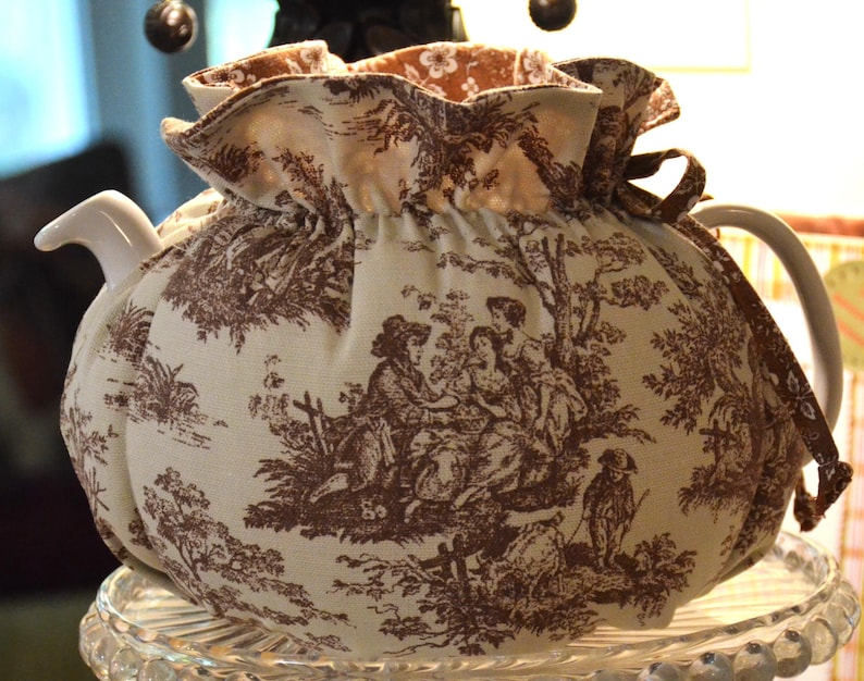 May include: A cream-colored teapot with a brown toile fabric cozy. The cozy features a pastoral scene with figures and trees. The teapot has a white handle and spout. The cozy is gathered at the top with a brown ribbon.