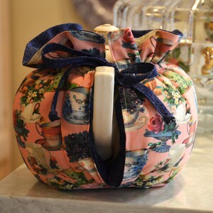 Puede incluir: Un calentador de t&eacute; rosa con un estampado floral con tazas de t&eacute; y flores. Tiene un cord&oacute;n azul marino y un ribete. El calentador est&aacute; dise&ntilde;ado para encajar alrededor de una tetera y mantener el t&eacute; caliente.