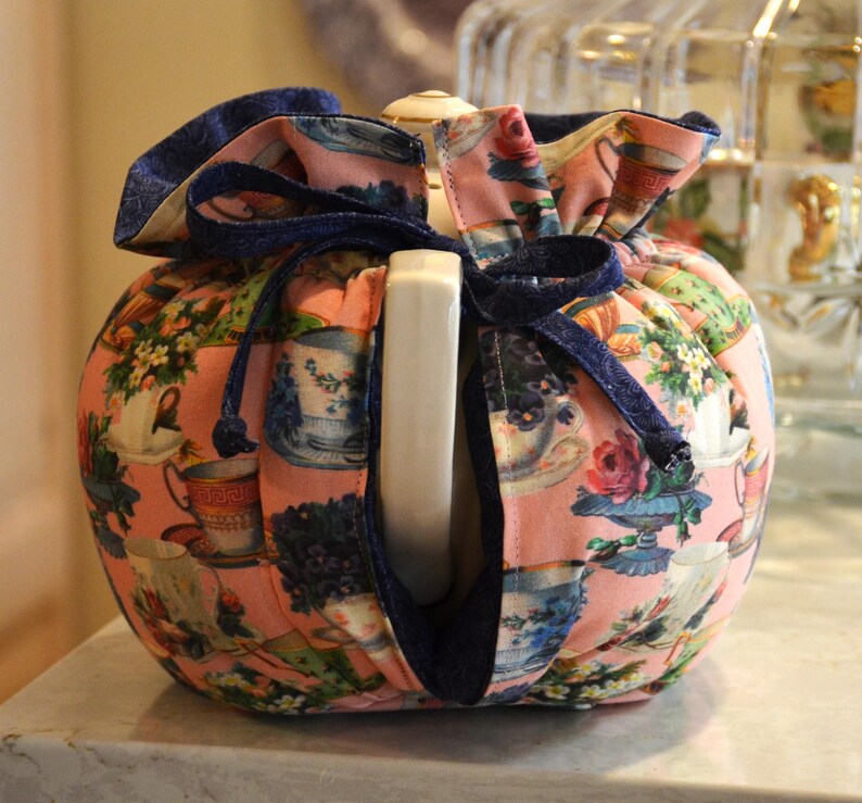 Puede incluir: Un calentador de t&eacute; rosa con un estampado floral con tazas de t&eacute; y flores. Tiene un cord&oacute;n azul marino y un ribete. El calentador est&aacute; dise&ntilde;ado para encajar alrededor de una tetera y mantener el t&eacute; caliente.