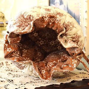 May include: A handmade drawstring bag with a floral and toile pattern in cream and brown. The bag's interior is a contrasting brown with white floral accents. The bag is open, revealing the interior, and sits on a lace doily.