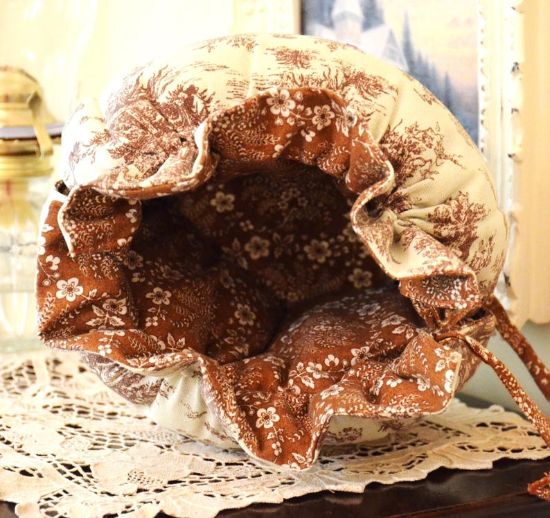 May include: A handmade drawstring bag with a floral and toile pattern in cream and brown. The bag's interior is a contrasting brown with white floral accents. The bag is open, revealing the interior, and sits on a lace doily.