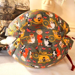 Puede incluir: Un art&iacute;culo de tela redondo, verde oliva, con un patr&oacute;n repetido de corgis, la Reina y banderas brit&aacute;nicas. El art&iacute;culo tiene un cierre de cord&oacute;n y es probablemente un contenedor o una cubierta.