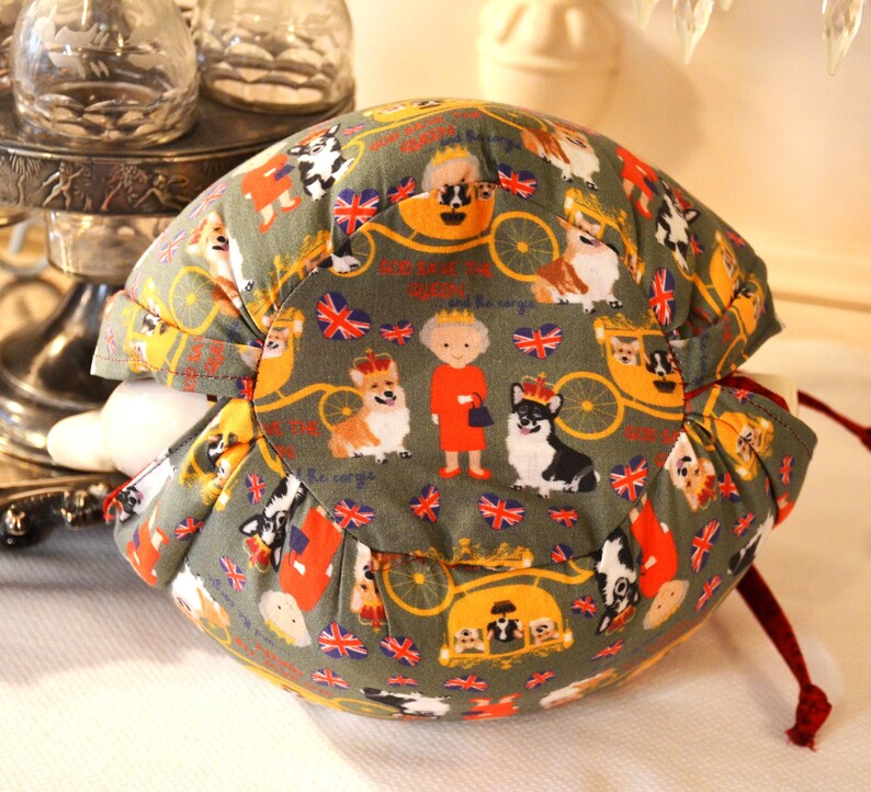 Puede incluir: Un art&iacute;culo de tela redondo, verde oliva, con un patr&oacute;n repetido de corgis, la Reina y banderas brit&aacute;nicas. El art&iacute;culo tiene un cierre de cord&oacute;n y es probablemente un contenedor o una cubierta.