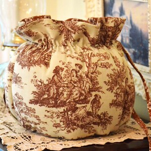 May include: A round, beige drawstring bag with a brown toile pattern depicting figures in a landscape. The bag has a ruffled top and a brown drawstring. The bag is sitting on a white lace doily.
