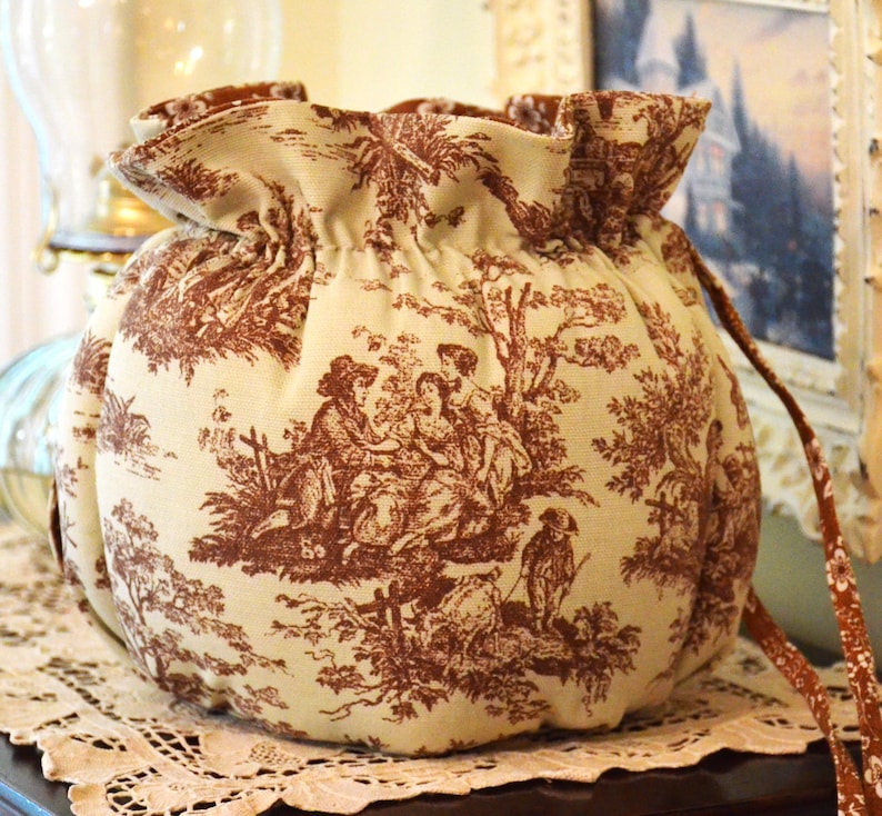 May include: A round, beige drawstring bag with a brown toile pattern depicting figures in a landscape. The bag has a ruffled top and a brown drawstring. The bag is sitting on a white lace doily.