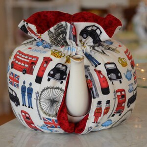 K&ouml;nnte beinhalten: Eine wei&szlig;e Teekannenhaube mit rotem Innenfutter und einem London-Themen-Druck. Der Druck zeigt rote Doppeldeckerbusse, schwarze Taxis, den Union Jack und andere ikonische Londoner Bilder. Die Haube ist f&uuml;r eine Teekanne konzipiert.