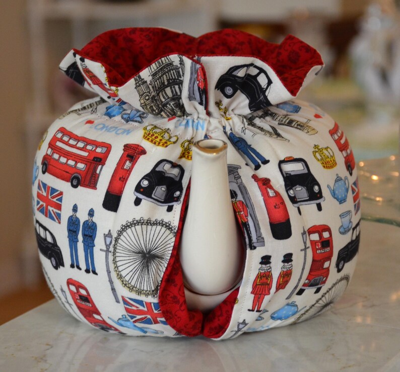 K&ouml;nnte beinhalten: Eine wei&szlig;e Teekannenhaube mit rotem Innenfutter und einem London-Themen-Druck. Der Druck zeigt rote Doppeldeckerbusse, schwarze Taxis, den Union Jack und andere ikonische Londoner Bilder. Die Haube ist f&uuml;r eine Teekanne konzipiert.