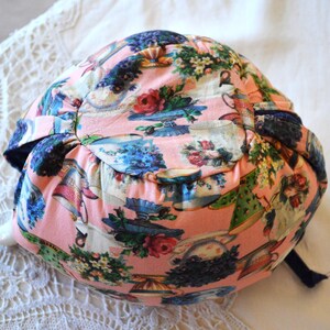 Puede incluir: Un sombrero de tela rosa con un estampado floral y de tazas de t&eacute;. El sombrero presenta un ala azul oscuro y una cinta a juego. El dise&ntilde;o incluye rosas, tazas de t&eacute; y arreglos florales en tonos rosas, azules y verdes.