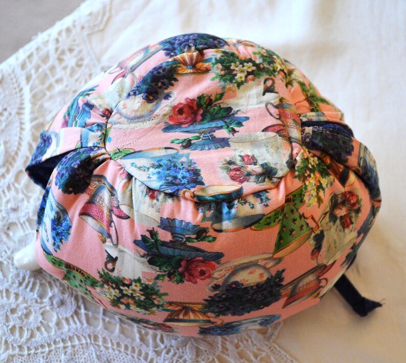 Puede incluir: Un sombrero de tela rosa con un estampado floral y de tazas de t&eacute;. El sombrero presenta un ala azul oscuro y una cinta a juego. El dise&ntilde;o incluye rosas, tazas de t&eacute; y arreglos florales en tonos rosas, azules y verdes.