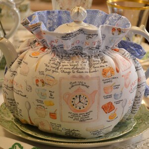 Może przedstawiać: Biały ceramiczny czajnik z materiałową osłoną na herbatę. Osłona jest ozdobiona ilustracjami filiżanek, bułeczek i tekstu. Czajnik stoi na zielono-białym talerzu.