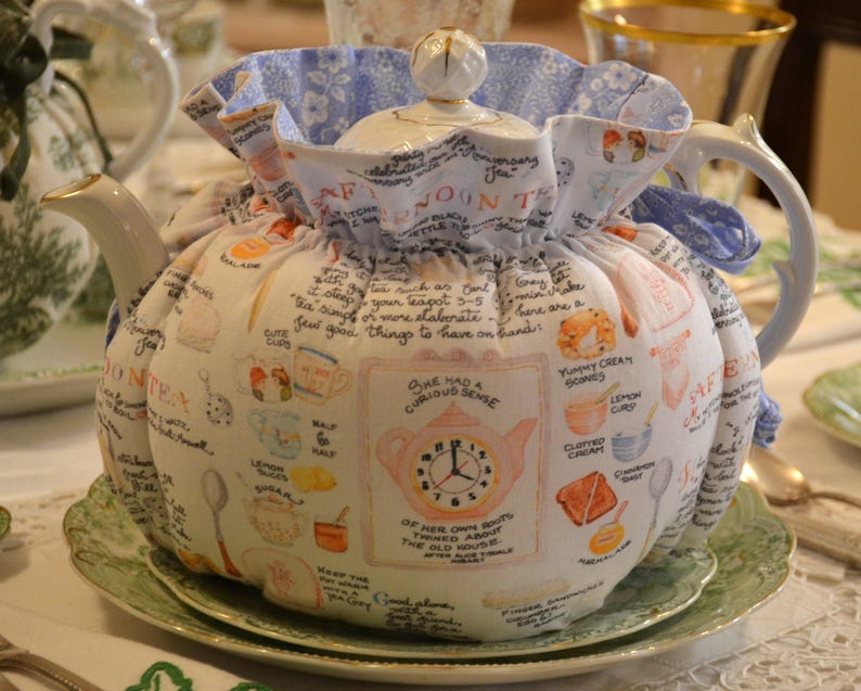 Może przedstawiać: Biały ceramiczny czajnik z materiałową osłoną na herbatę. Osłona jest ozdobiona ilustracjami filiżanek, bułeczek i tekstu. Czajnik stoi na zielono-białym talerzu.