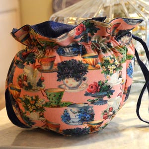 Puede incluir: Una bolsa rosa con cord&oacute;n y un estampado floral de tazas de t&eacute;. El exterior de la bolsa presenta un dise&ntilde;o repetido de tazas de t&eacute;, platillos y arreglos florales en varios colores. El interior y el cord&oacute;n son de color azul marino.