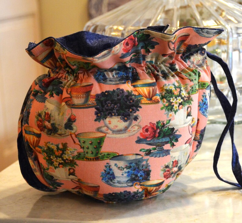 Puede incluir: Una bolsa rosa con cord&oacute;n y un estampado floral de tazas de t&eacute;. El exterior de la bolsa presenta un dise&ntilde;o repetido de tazas de t&eacute;, platillos y arreglos florales en varios colores. El interior y el cord&oacute;n son de color azul marino.