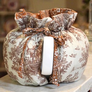 May include: A white ceramic teapot with a brown and white toile fabric cozy. The fabric features a pastoral scene with figures, trees, and animals. The cozy is tied with a brown ribbon.