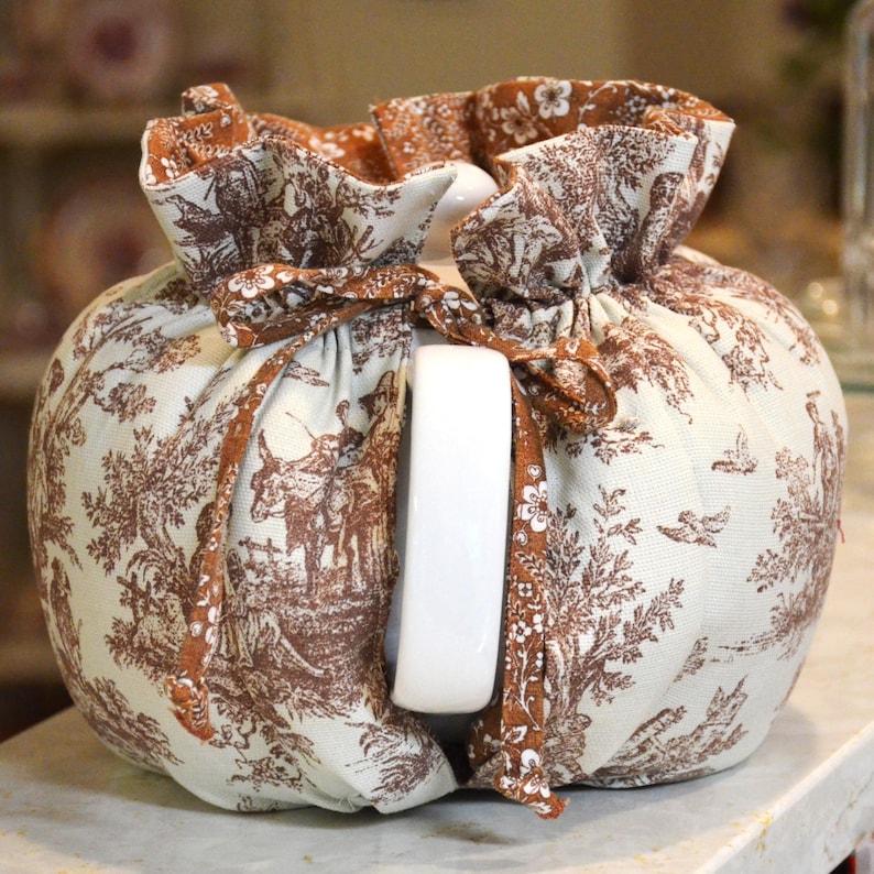 May include: A white ceramic teapot with a brown and white toile fabric cozy. The fabric features a pastoral scene with figures, trees, and animals. The cozy is tied with a brown ribbon.