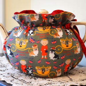 Puede incluir: Un cubreteiera decorativo con un estampado caprichoso con corgis, una figura real y un carruaje sobre un fondo verde. El cubreteiera tiene un ribete de terciopelo rojo y un cierre de cord&oacute;n. La tetera tiene un asa y un pico dorados.