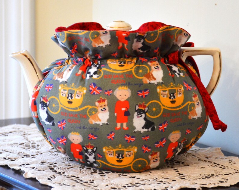 Puede incluir: Un cubreteiera decorativo con un estampado caprichoso con corgis, una figura real y un carruaje sobre un fondo verde. El cubreteiera tiene un ribete de terciopelo rojo y un cierre de cord&oacute;n. La tetera tiene un asa y un pico dorados.