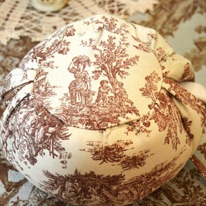 May include: A round, beige fabric pouch with a brown toile pattern depicting figures in a pastoral scene. The pouch is gathered at the top with a drawstring. The fabric has a soft, textured appearance.