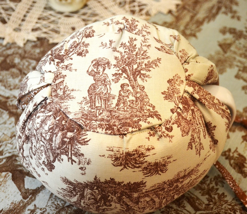 May include: A round, beige fabric pouch with a brown toile pattern depicting figures in a pastoral scene. The pouch is gathered at the top with a drawstring. The fabric has a soft, textured appearance.
