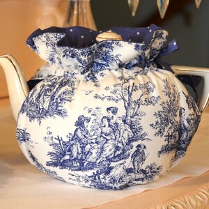Op de afbeelding: Een witte porseleinen theepot met een blauw-wit toile de Jouy-patroon, bedekt met een bijpassende theemuts. De theemuts is aan de bovenkant verzameld met een blauw lint. De theepot heeft een gouden handvat en tuit.