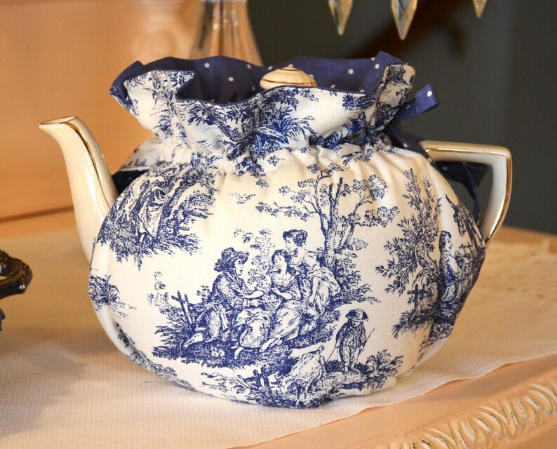 Op de afbeelding: Een witte porseleinen theepot met een blauw-wit toile de Jouy-patroon, bedekt met een bijpassende theemuts. De theemuts is aan de bovenkant verzameld met een blauw lint. De theepot heeft een gouden handvat en tuit.