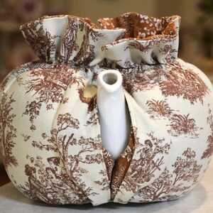 May include: A white ceramic teapot peeks out from a fabric tea cozy. The cozy is cream-colored with a brown toile pattern depicting scenes of people and trees. The top of the cozy has a ruffled edge.