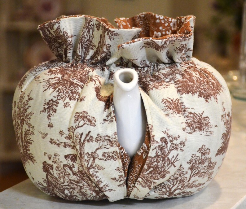 May include: A white ceramic teapot peeks out from a fabric tea cozy. The cozy is cream-colored with a brown toile pattern depicting scenes of people and trees. The top of the cozy has a ruffled edge.