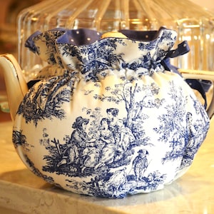 May include: A white and blue toile teapot cozy. The cozy features a blue and white pastoral scene with figures and trees. The top is blue with white polka dots and a blue ribbon. The teapot has a gold handle and spout.