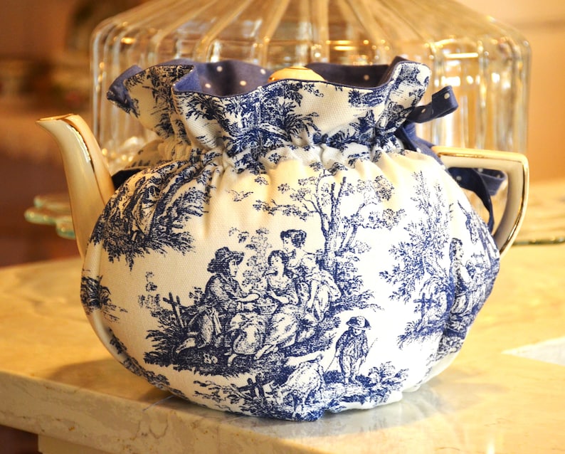 Op de afbeelding: Een wit en blauw toile de Jouy theepot hoes. De hoes heeft een blauw-witte pastorale sc&egrave;ne met figuren en bomen. De bovenkant is blauw met witte stippen en een blauw lint. De theepot heeft een gouden handvat en tuit.