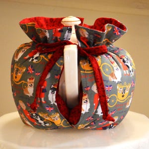 Puede incluir: Un calentador de t&eacute; gris con interior rojo y lazo rojo. El tejido presenta im&aacute;genes de perros corgi, coronas y carruajes. Envuelve una tetera blanca.